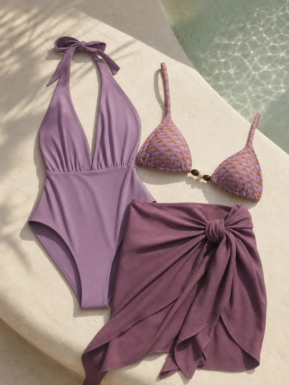 A lilac one-piece, patterned swim top, and plum resort wrap beside a pool.