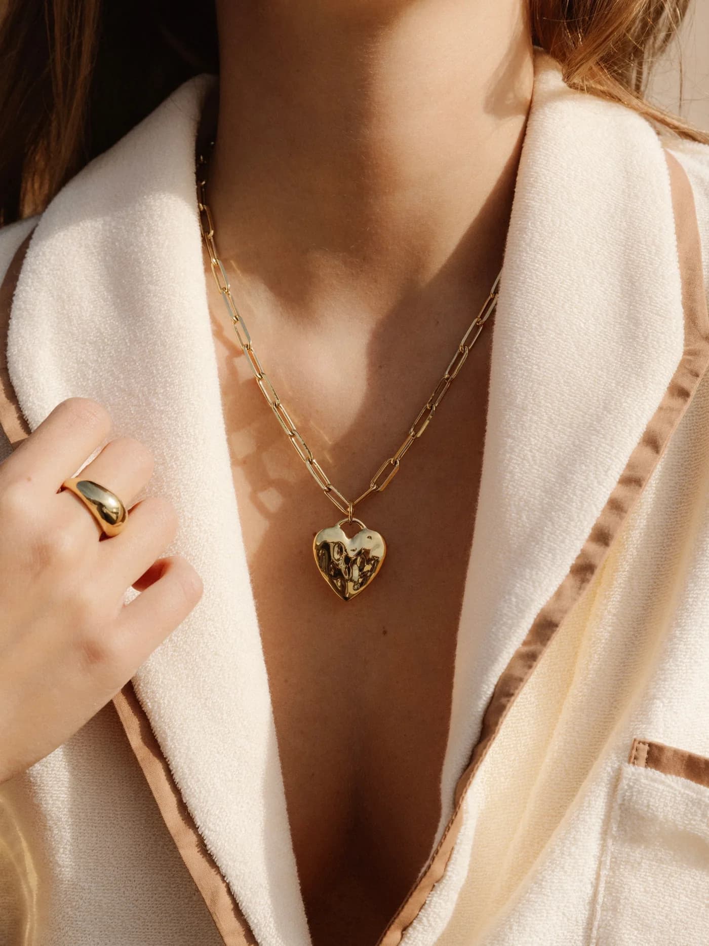 A James Michelle heart locket and layered gold chains worn with a cream jacket.