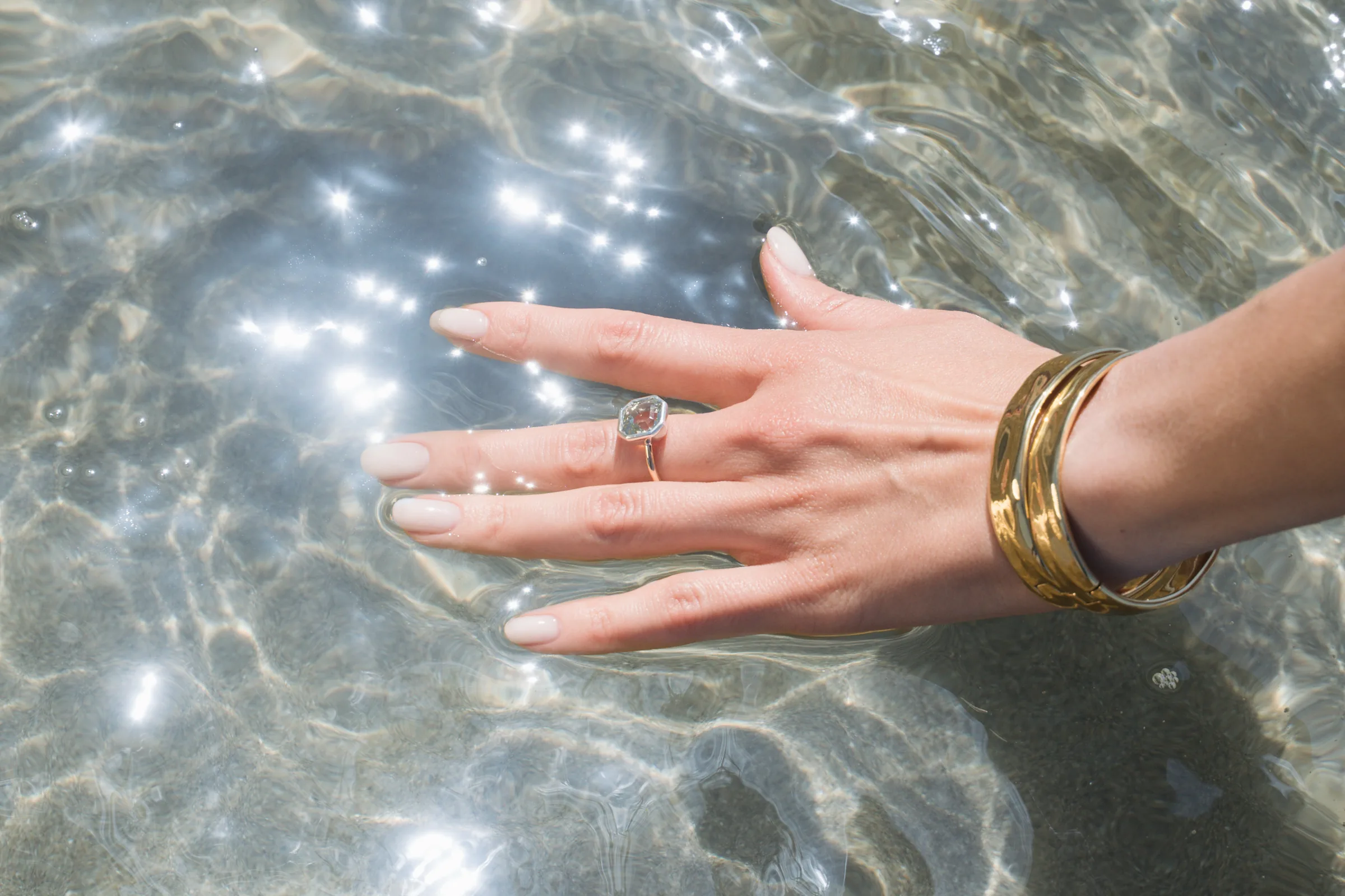 A hand wearing James Michelle rings and stacked gold bangles resting in sunlit water.