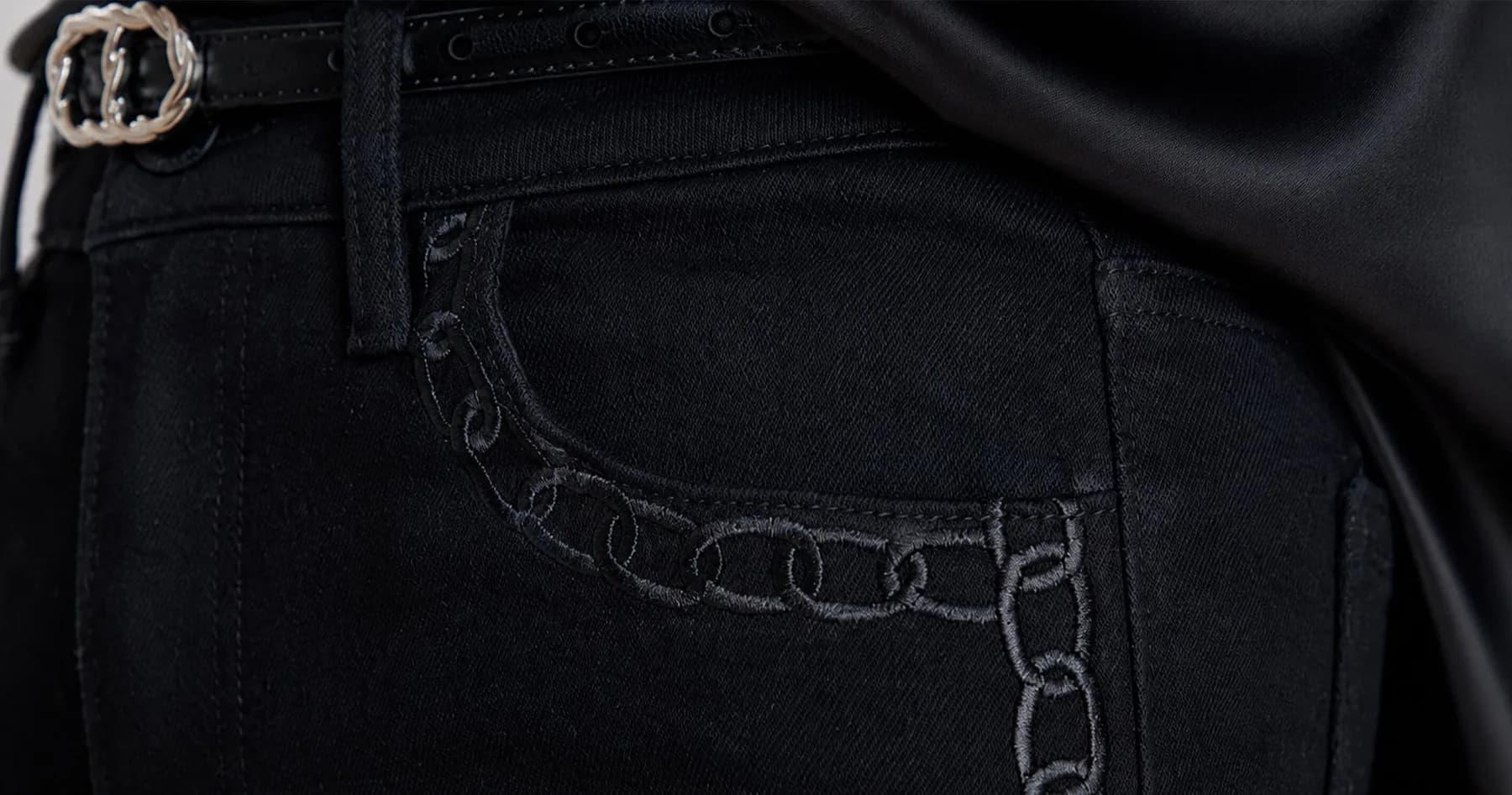 A close-up of Noli black denim showing the chain-link pocket hardware and topstitching.